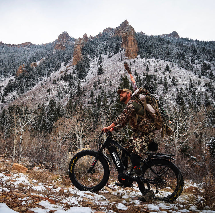 Hunter in camouflage gear with a rifle riding an electric bike on a wooded trail, navigating rugged terrain for a stealthy approach