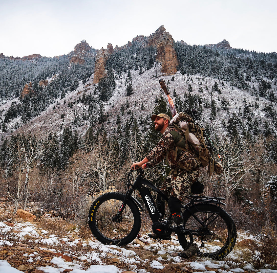 Hunter in camouflage gear with a rifle riding an electric bike on a wooded trail, navigating rugged terrain for a stealthy approach